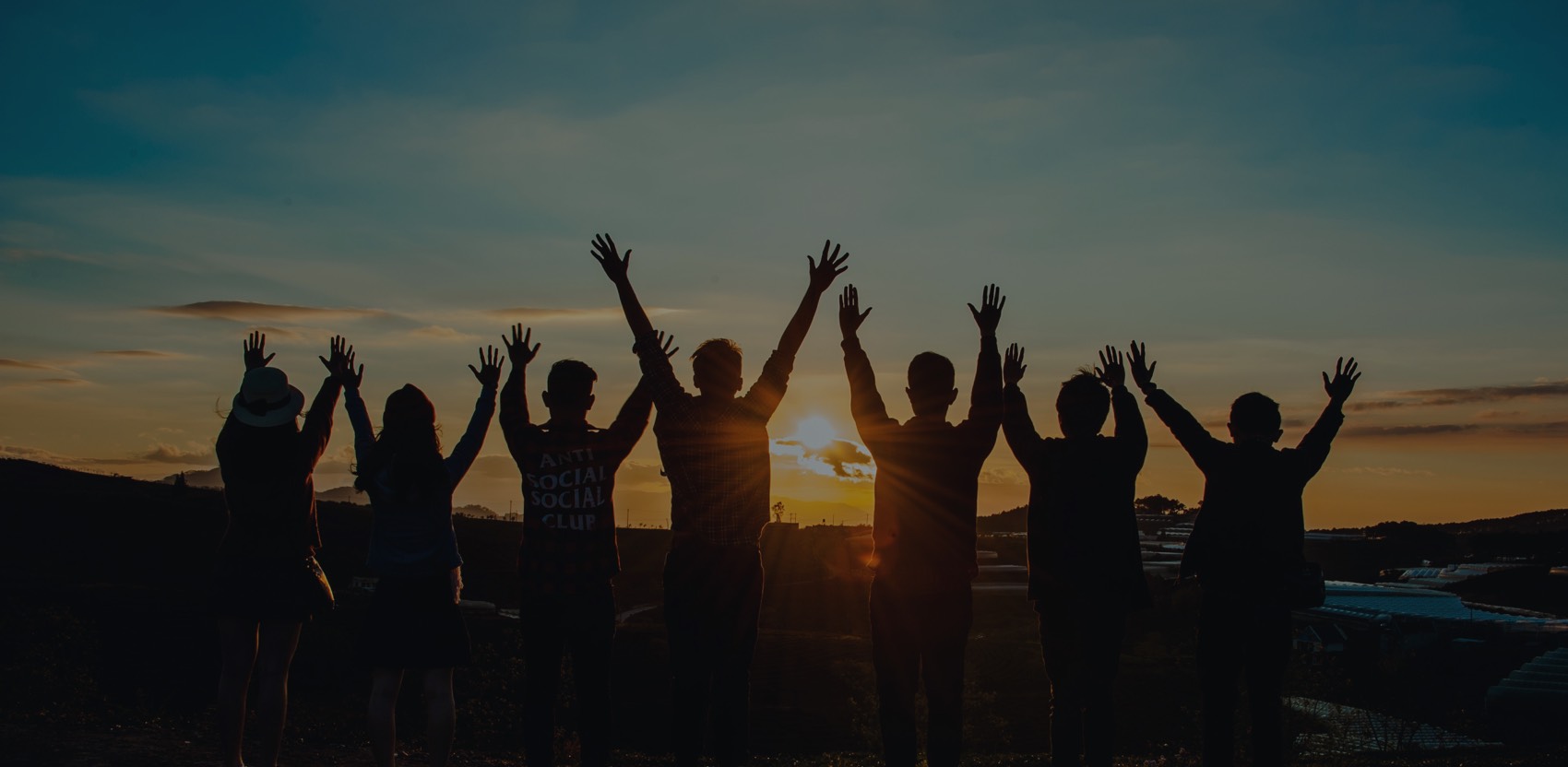 Silhouettes of a group of people with raised hands standing together at sunset, symbolizing unity and collaboration.